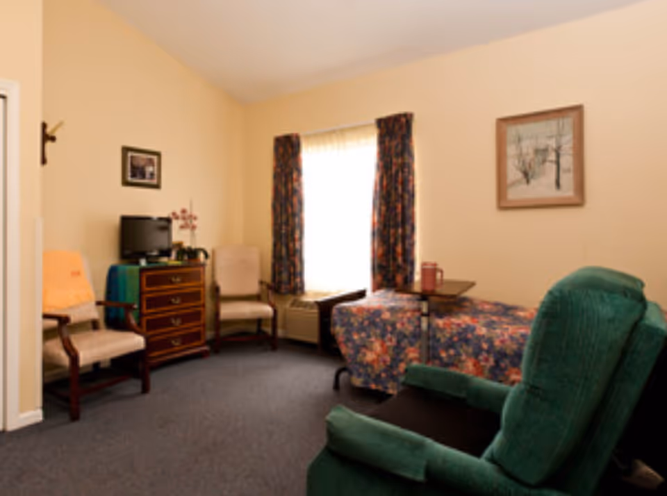 A cozy room with beige walls and carpeted floor featuring a floral-patterned bed, a green recliner chair, two beige armchairs, a wooden dresser with a small TV on top, a window with floral curtains, and framed artwork on the walls.
