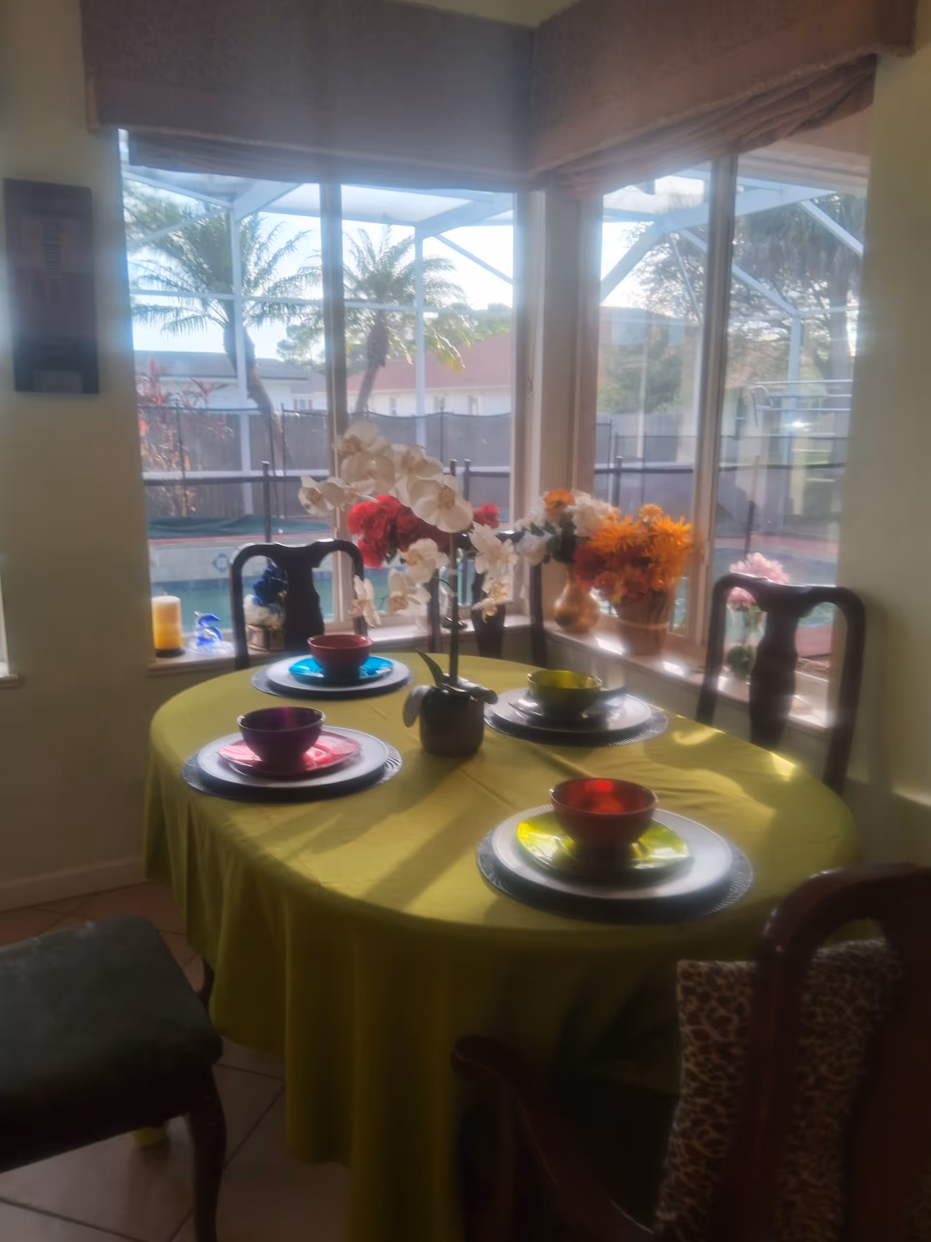 Round dining table set for four with colorful bowls and plates and floral centerpieces in front of large windows overlooking a screened backyard pool.