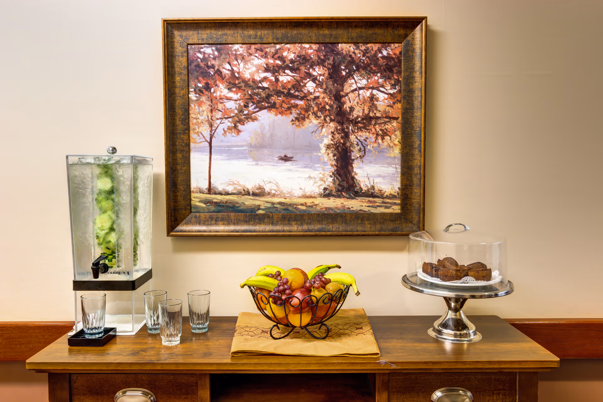 A wooden sideboard with a glass water dispenser filled with cucumber slices and four empty glasses on the left, a bowl of assorted fresh fruit including bananas, grapes, and oranges in the center, and a silver cake stand with a glass cover containing muffins on the right. Above the sideboard hangs a framed painting of a tree by a lake with a person in a small boat in the background.
