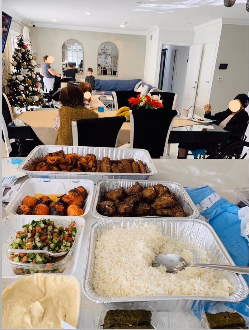 A dining area in a senior living facility with a table set with trays of food including rice, grilled meats, roasted vegetables, salad, and hummus. In the background, several people are seated and standing near a decorated Christmas tree and a couch with mirrors on the wall.