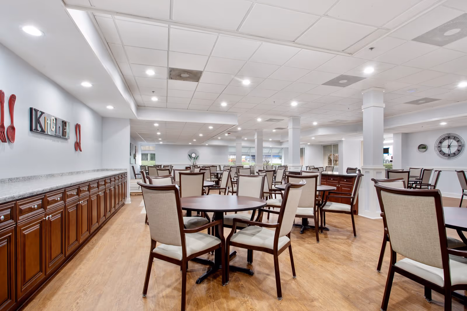 A spacious dining room with multiple round wooden tables and cushioned chairs arranged neatly on a wooden floor. The room has a white ceiling with recessed lighting and white walls. On the left wall, there is a decorative sign spelling 'KITCHEN' with red utensils on either side. A large clock is visible on the right wall.