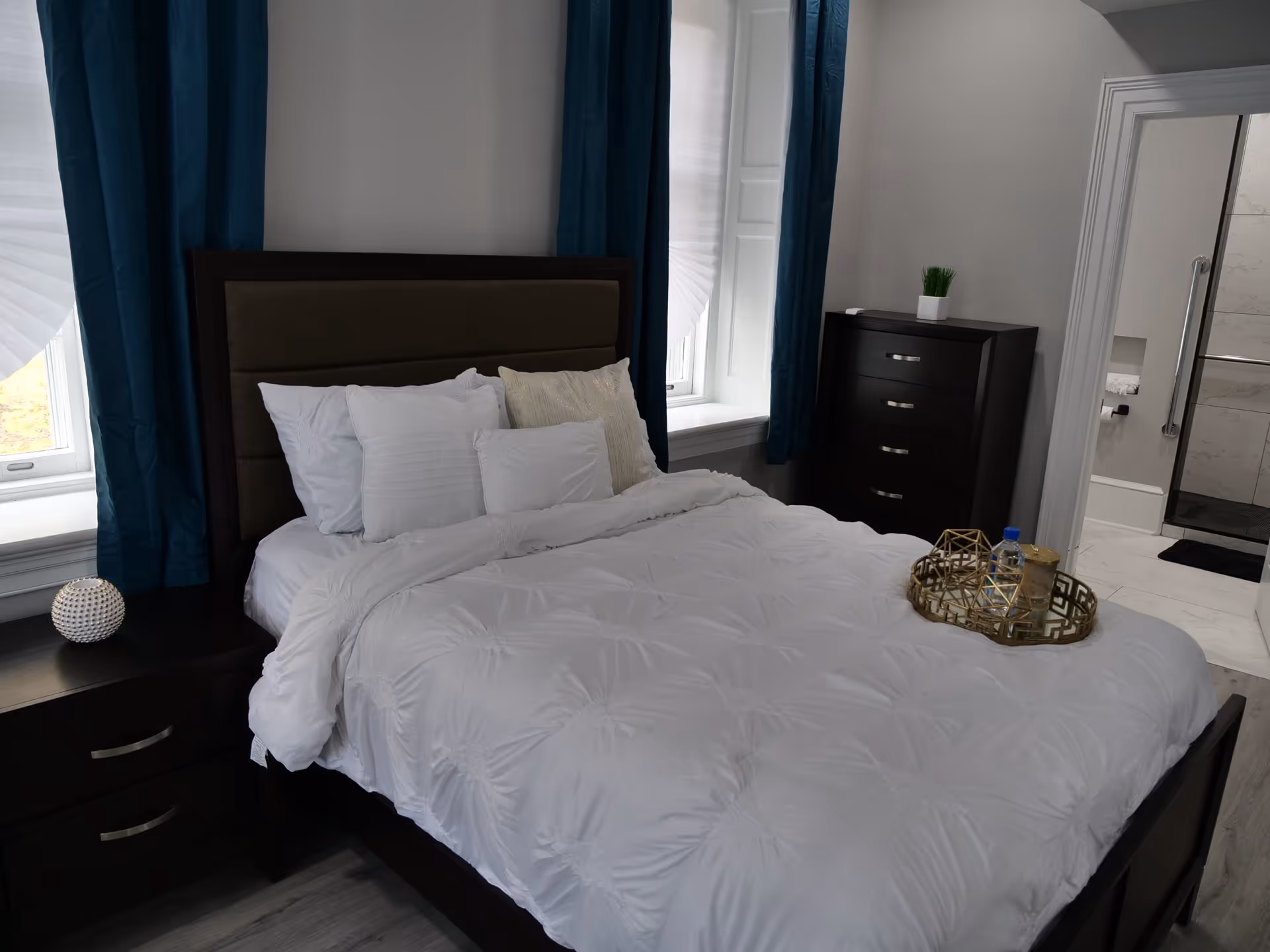 A neatly made bed with white bedding and multiple pillows in a bedroom. The bed has a dark wooden frame and headboard. There are two windows with blue curtains on either side of the bed. A dark wooden nightstand with a decorative item is on the left side of the bed. On the right side, there is a dark wooden dresser with a small plant on top. An open door reveals a bathroom with a shower and grab bars.