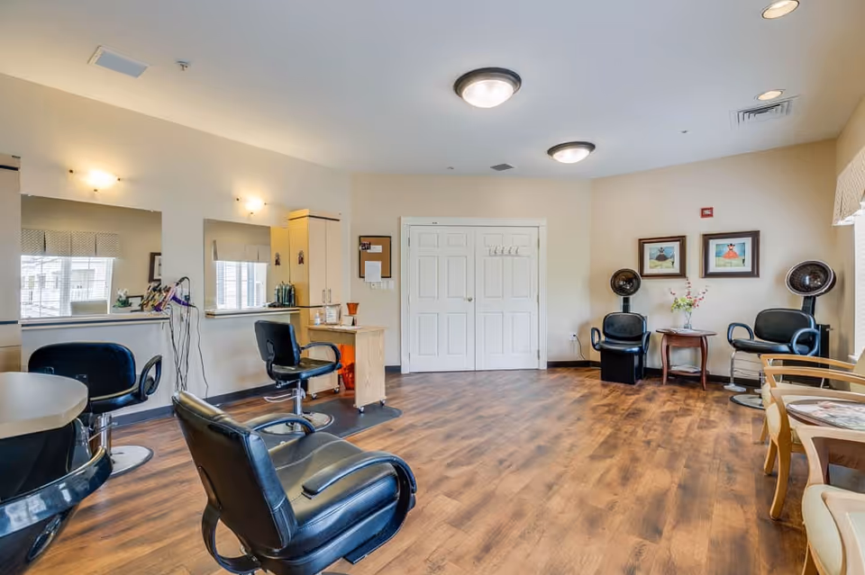 Interior of a salon area in a senior living facility with wooden flooring, black salon chairs, mirrors on the wall, hair dryers, and a small table with flowers and framed artwork on the walls.