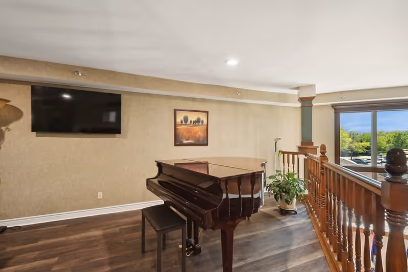 Interior view of a room with a grand piano and a piano bench on wooden flooring. The room has beige walls with a framed landscape painting, a mounted flat-screen TV, a potted plant near a wooden railing, and a large window showing greenery outside.