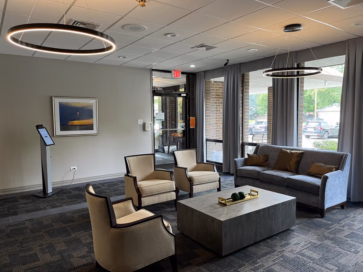 A modern seating area inside Tower Hill Healthcare Center featuring a gray sofa with brown pillows, three beige armchairs with dark wooden frames, and a rectangular coffee table with decorative items. The room has large windows with gray curtains, circular ceiling lights, and a framed picture on the wall near the entrance door.