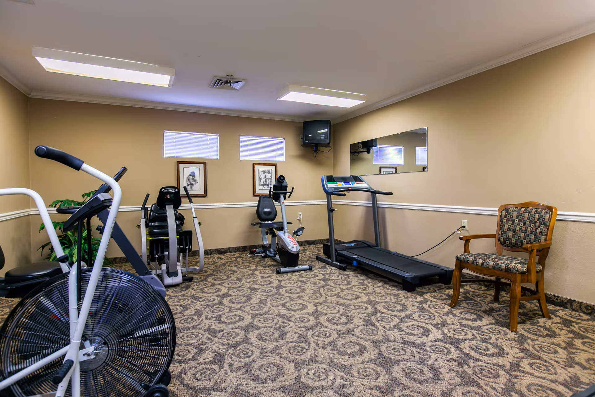 A small fitness room with exercise equipment including a stationary bike, rowing machine, recumbent bike, and treadmill. The room has beige walls, patterned carpet, two small windows with blinds, a wall-mounted TV, a large mirror, and a wooden chair with patterned upholstery.