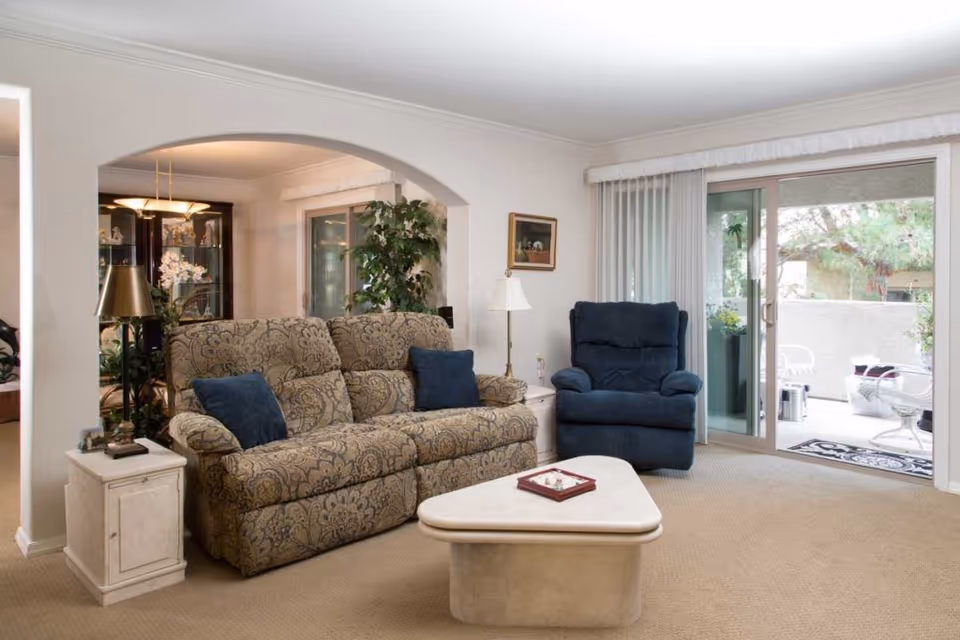 A cozy living room with a patterned beige sofa adorned with two blue cushions, a blue armchair, a white side table with a lamp, and a triangular coffee table with a decorative item on it. The room has beige carpet flooring, white walls, and a large sliding glass door leading to an outdoor patio with white chairs and greenery visible outside.