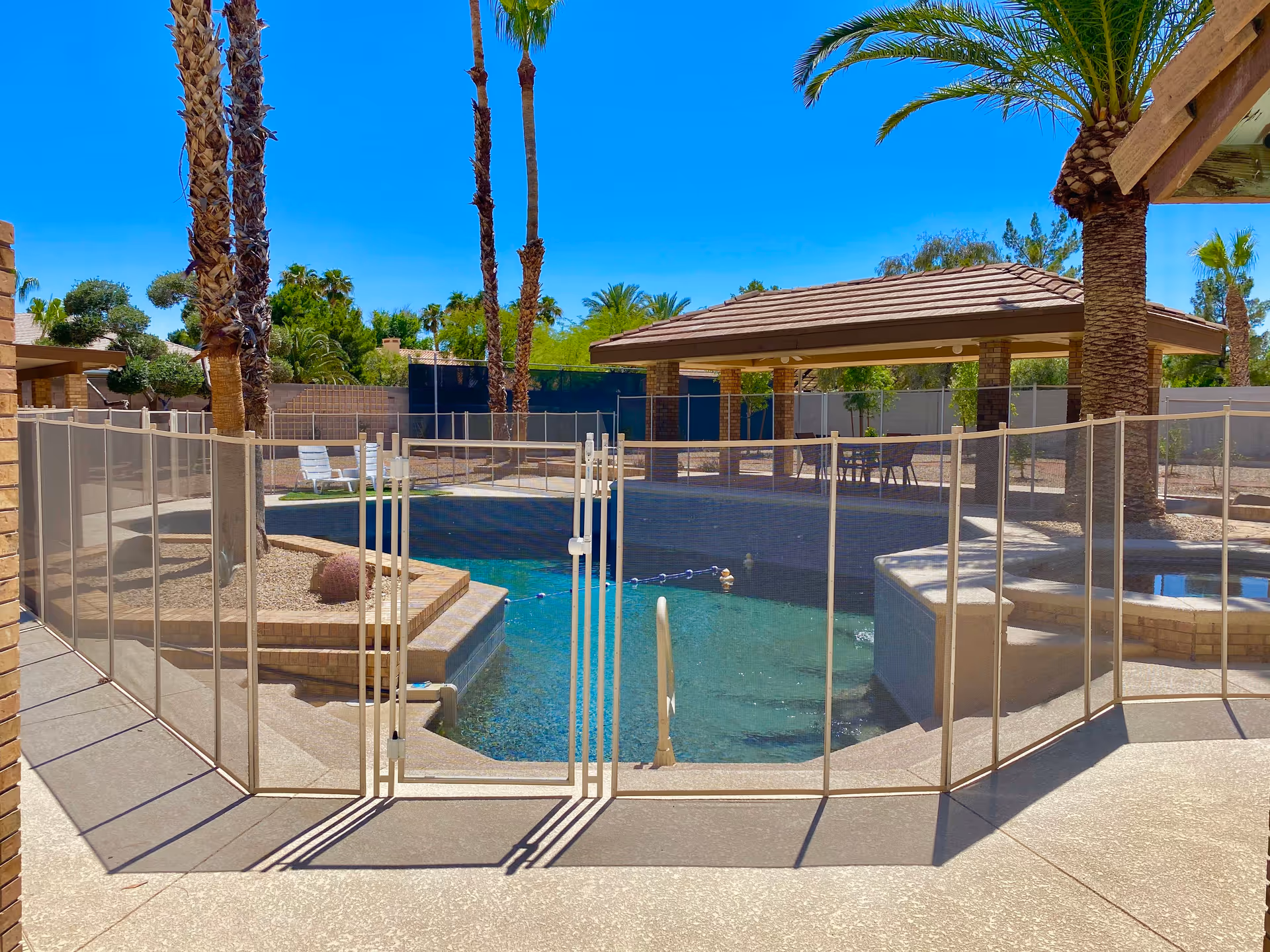 Outdoor swimming pool area surrounded by a safety fence with a gate. Palm trees and a covered seating area with tables and chairs are visible in the background under a clear blue sky.