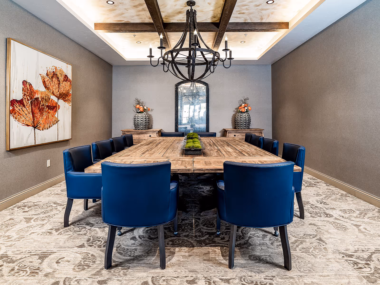 Modern dining room with a long wooden table surrounded by blue chairs, a chandelier overhead, and artwork on the wall.
