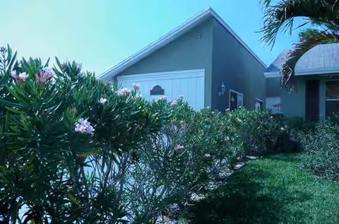 Exterior view of Orange Blossoms Villa with hedges, pink flowers, and a gray building.