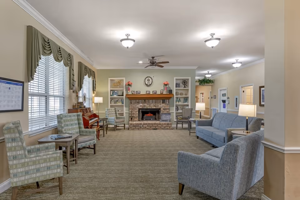 A cozy living room area in a senior living facility with a brick fireplace centered on the far wall, flanked by built-in shelves decorated with framed photos, vases, and flowers. The room has multiple seating options including patterned armchairs, a blue sofa, and a piano against the left wall under windows with green valances. Several lamps provide warm lighting and ceiling fans are mounted on the ceiling.
