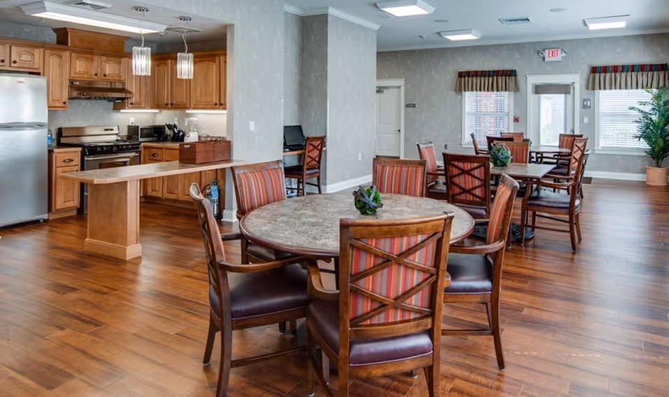 A spacious dining area with multiple round tables and wooden chairs with striped cushions. The room features a kitchen area with wooden cabinets, a stainless steel refrigerator, stove, and a long wooden counter. The floor is wooden, and there are windows with blinds and valances, as well as an exit door in the background.
