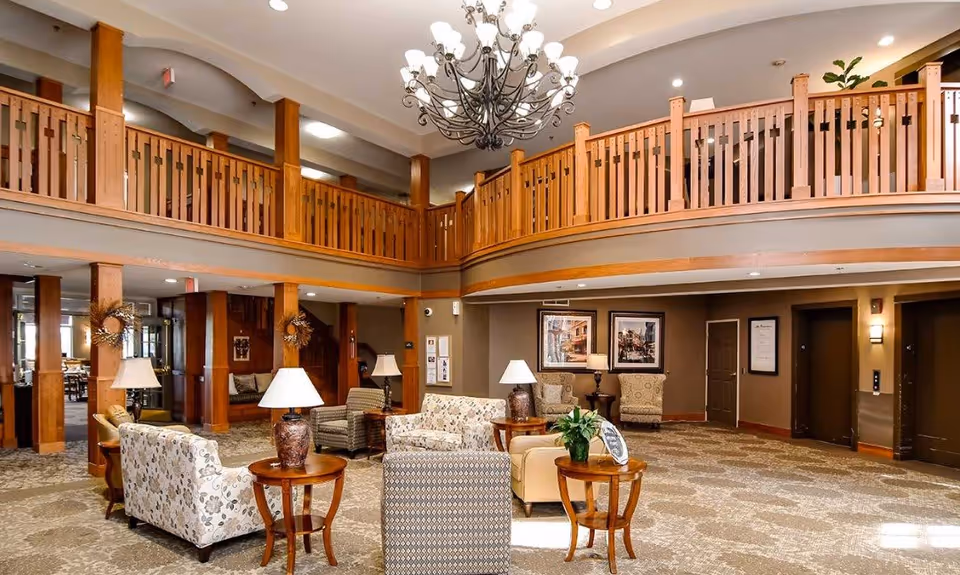 Spacious senior living facility common area with comfortable armchairs and sofas arranged around wooden side tables with lamps and plants. The room features a high ceiling with a large decorative chandelier and a wooden balcony railing on the upper level. The walls are adorned with framed artwork, and there are multiple doorways and an elevator visible in the background.