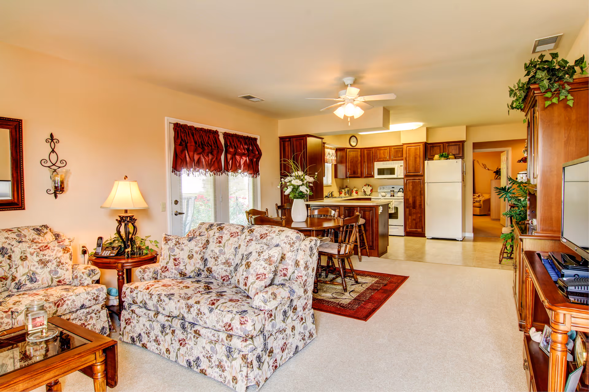 A cozy living room and kitchen area in a senior living facility. The living room features floral patterned sofas, a wooden coffee table, a side table with a lamp, and a TV on a wooden stand. The kitchen has wooden cabinets, a white refrigerator, a microwave, and a dining table with chairs on a red rug. The room is well-lit with a ceiling fan light and natural light coming through a window with red curtains.