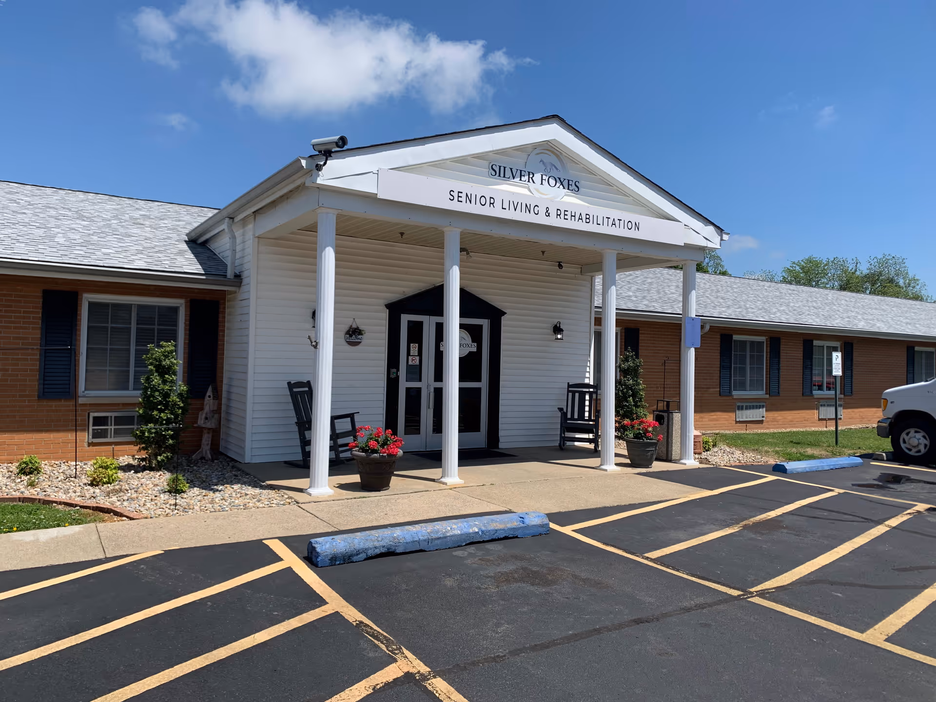 Front entrance of Silver Foxes Senior Living & Rehabilitation with a covered portico, columns, parking spaces, and potted plants.
