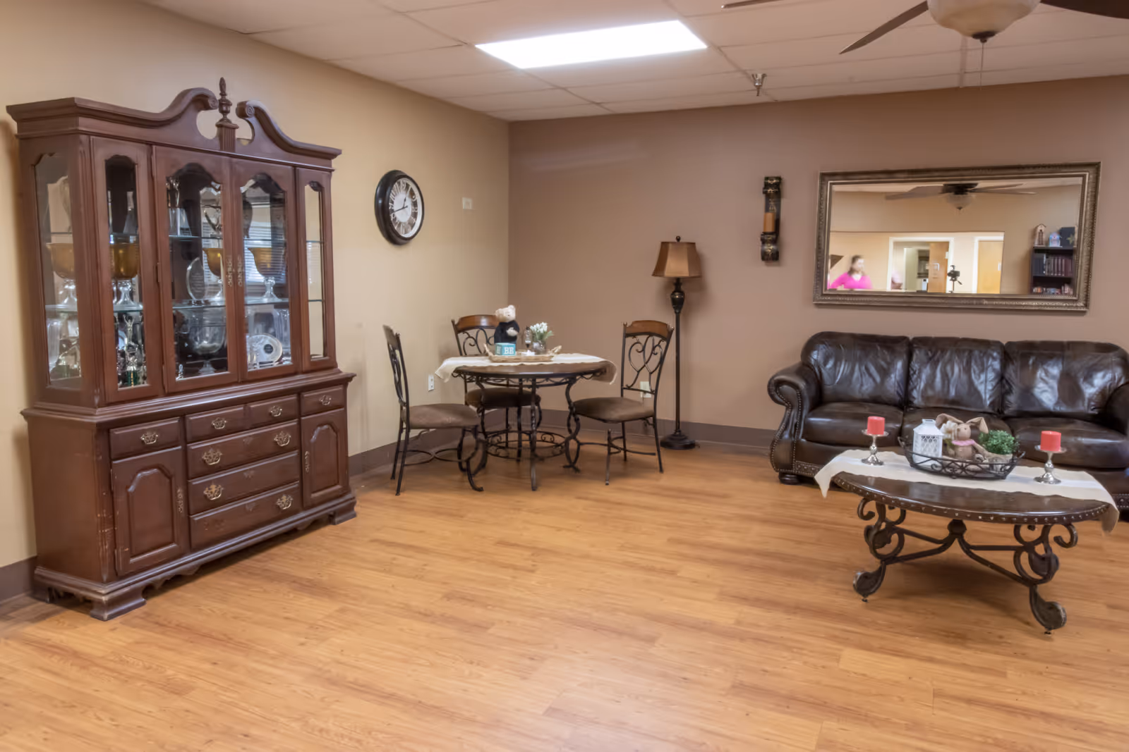 A cozy living room area with a wooden china cabinet filled with glassware on the left, a small round table with three chairs in the center against a beige wall, and a dark brown leather sofa on the right. A decorative coffee table with candles and a small plant is in front of the sofa. A large mirror hangs on the wall above the sofa, reflecting part of the room and a person in a pink shirt. The floor is wooden, and the ceiling has fluorescent lighting and a ceiling fan.