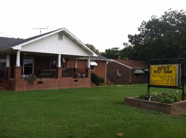 Exterior view of Boyd's Rest Home, Inc., a single-story brick building with a covered porch and a well-maintained lawn. A yellow sign in front of the building displays the facility's name and contact information.