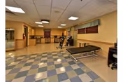 A spacious rehabilitation room with checkered blue and beige flooring, a treatment table, exercise equipment, and a desk with a chair. The room has beige walls with wooden trim and windows with brown blinds and green valances.