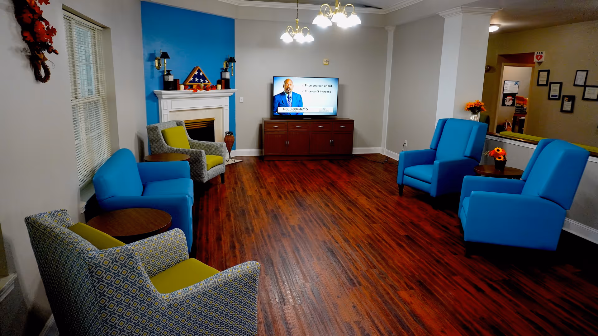A cozy living room with wooden flooring, featuring four blue armchairs and two patterned armchairs with green cushions arranged around small round wooden tables. A white fireplace with a folded American flag in a triangular display case sits against a blue accent wall. A flat-screen TV is mounted on a wooden cabinet, and there are two ceiling light fixtures with multiple bulbs. The room has windows with blinds and some decorative flowers on tables.