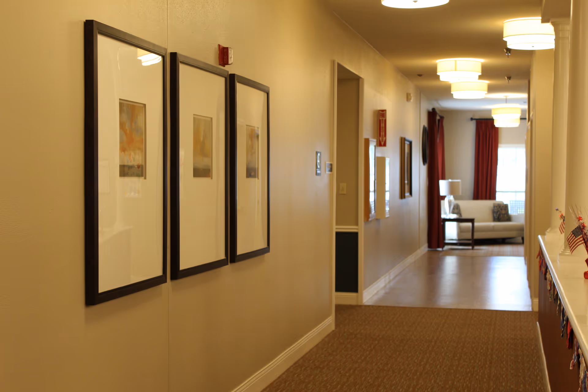 Well-lit hallway in an assisted living facility with three framed pictures on the left wall and a seating area with a sofa and lamp visible at the far end.