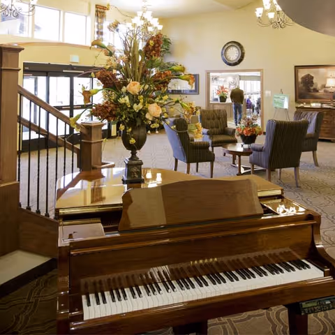 Interior view of a senior living facility lounge area featuring a polished Yamaha grand piano in the foreground, a large floral arrangement on a table behind it, several upholstered chairs arranged around a coffee table, a wall clock, and a person walking in the background near a doorway.