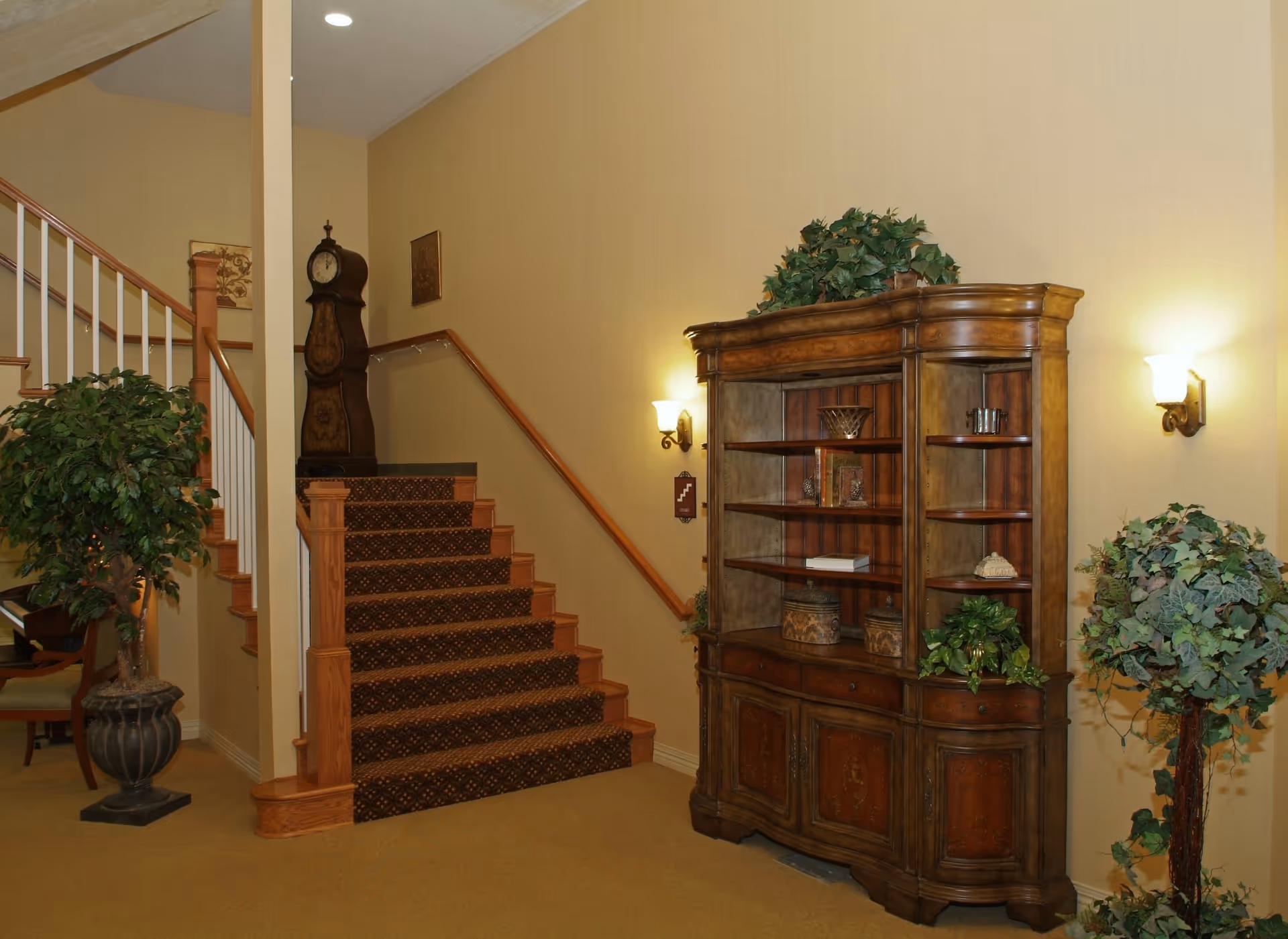 Interior view of a senior living community featuring a carpeted staircase with wooden railings, a large wooden cabinet with shelves displaying decorative items, two wall-mounted lamps, and two potted plants on either side of the cabinet.