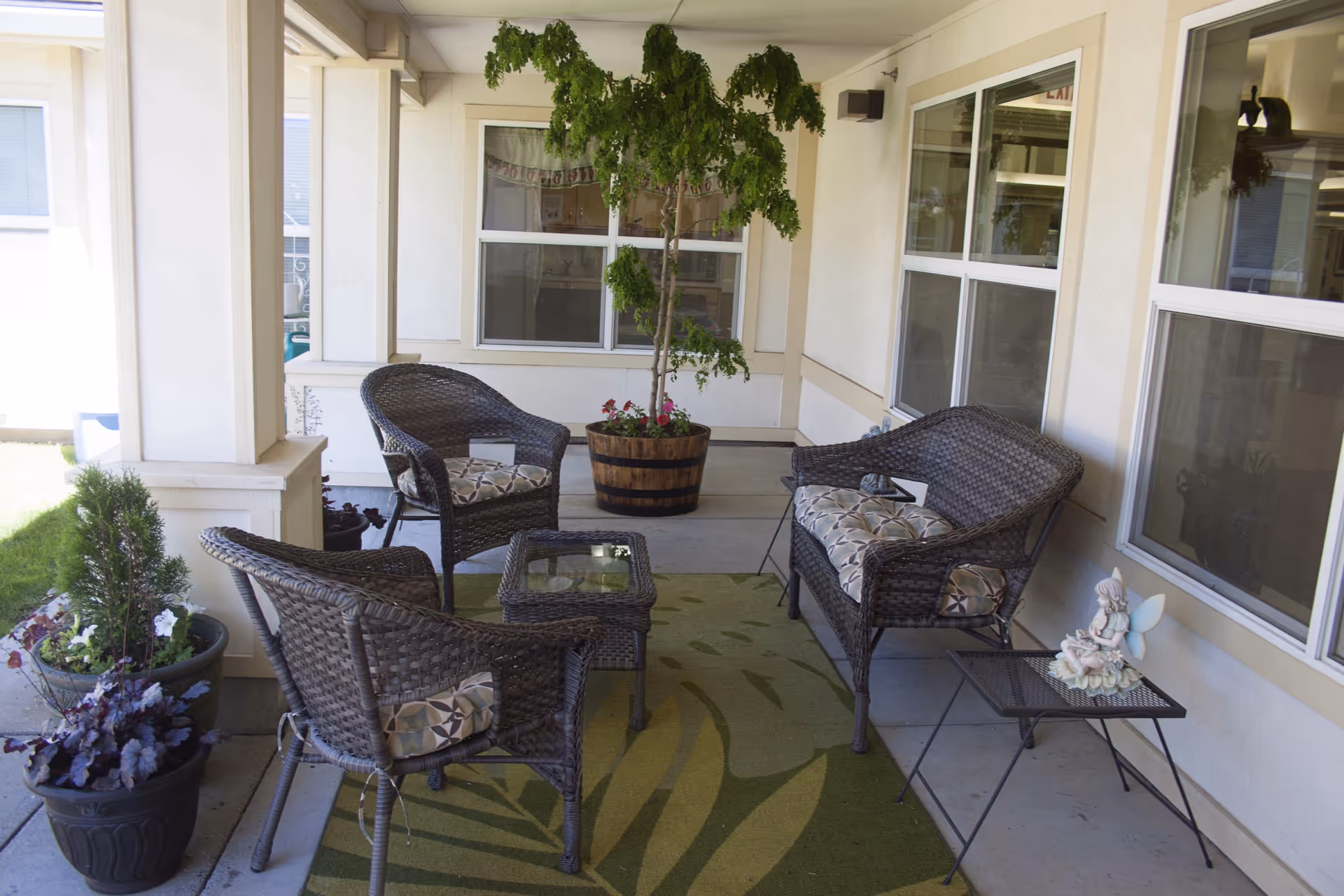 Outdoor covered patio area with wicker furniture including two armchairs, a loveseat, and a small glass-top table. Cushions with patterned fabric are on the chairs. There are potted plants and a small tree in a wooden barrel planter. A small side table holds a fairy figurine. The area is adjacent to a building with multiple windows.