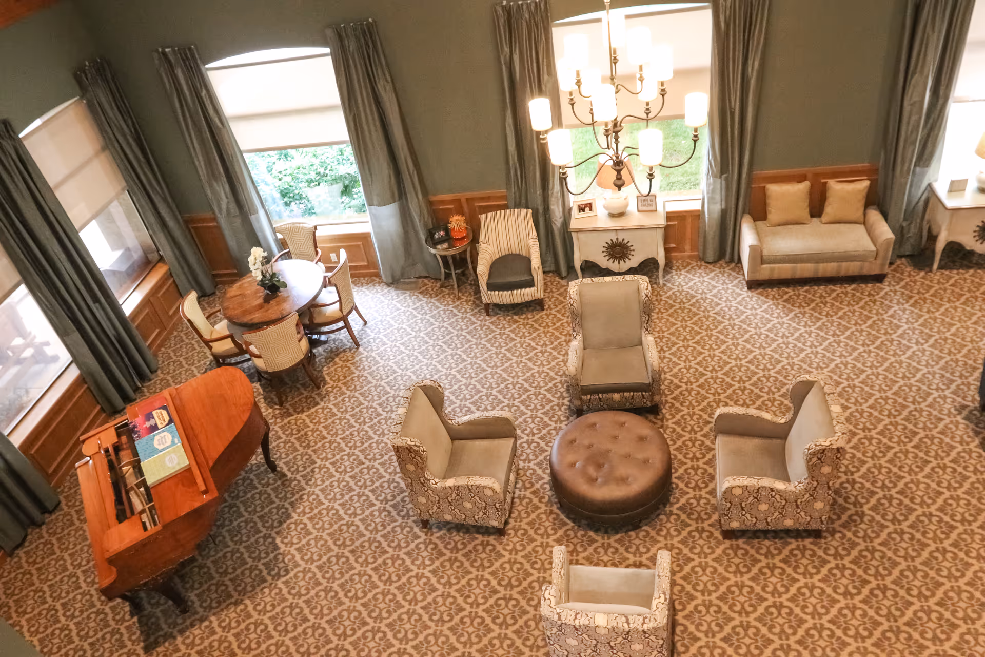 Aerial view of a furnished senior living common room with armchairs arranged around a round ottoman, a grand piano, a small dining table, and large windows with drapes.