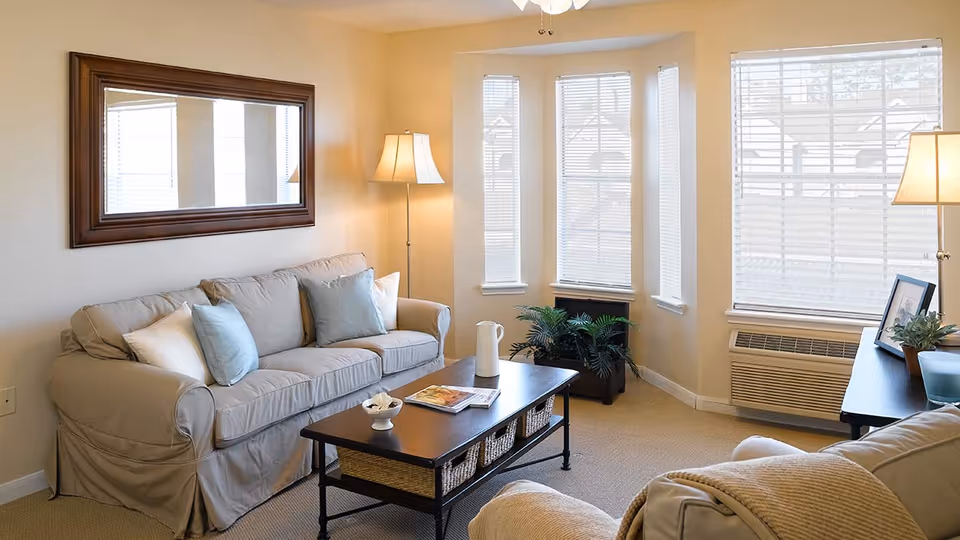 A cozy living room with a beige sofa adorned with light blue and white pillows, a dark wooden coffee table with baskets underneath, a floor lamp, a large wall mirror, and three windows with blinds letting in natural light. There is also a small table with a lamp and framed photos, and a potted plant near the windows.