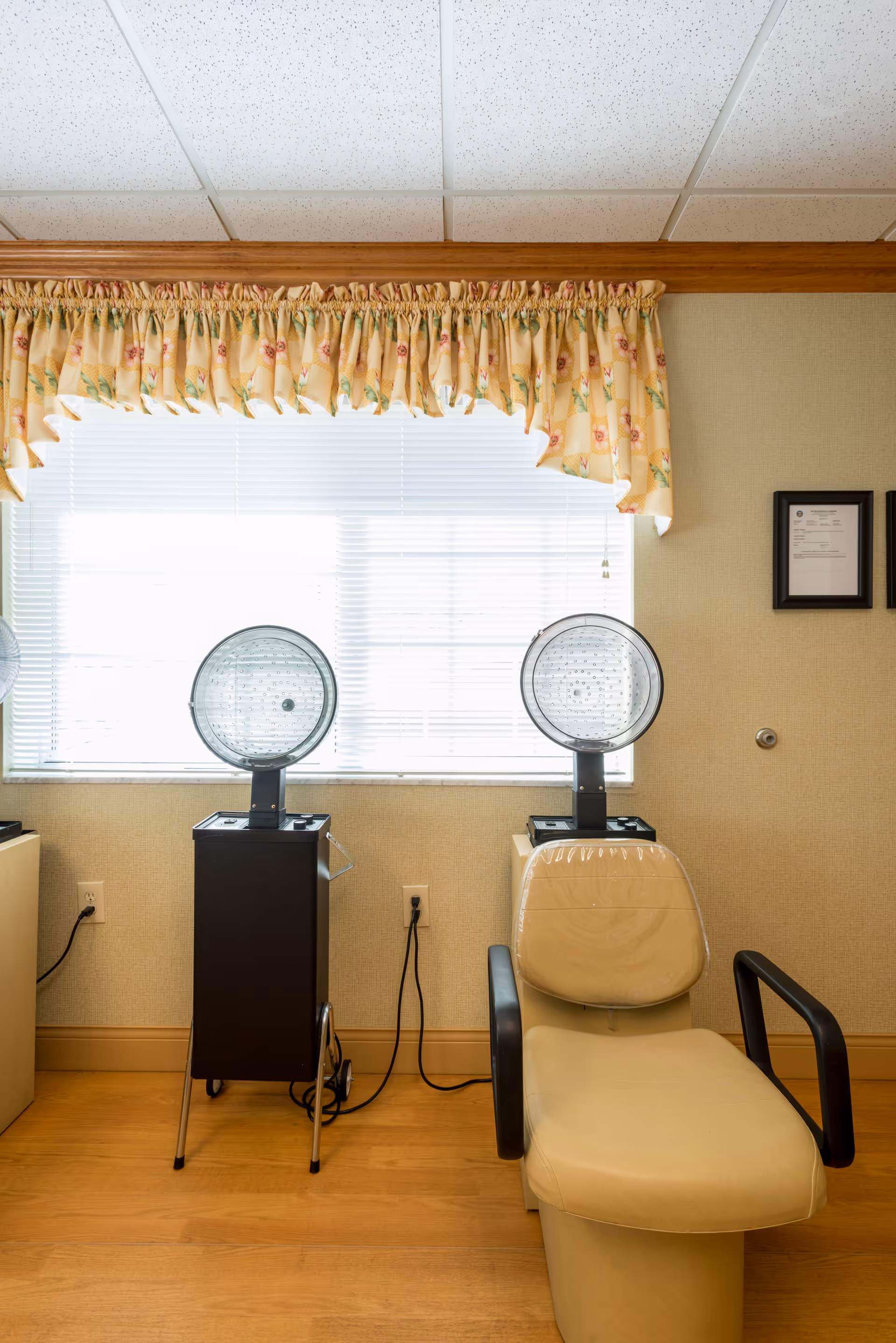 A treatment room with a beige reclining chair covered in plastic, two vintage hair dryer machines on stands, a window with blinds and floral valance, and framed certificates on the wall.