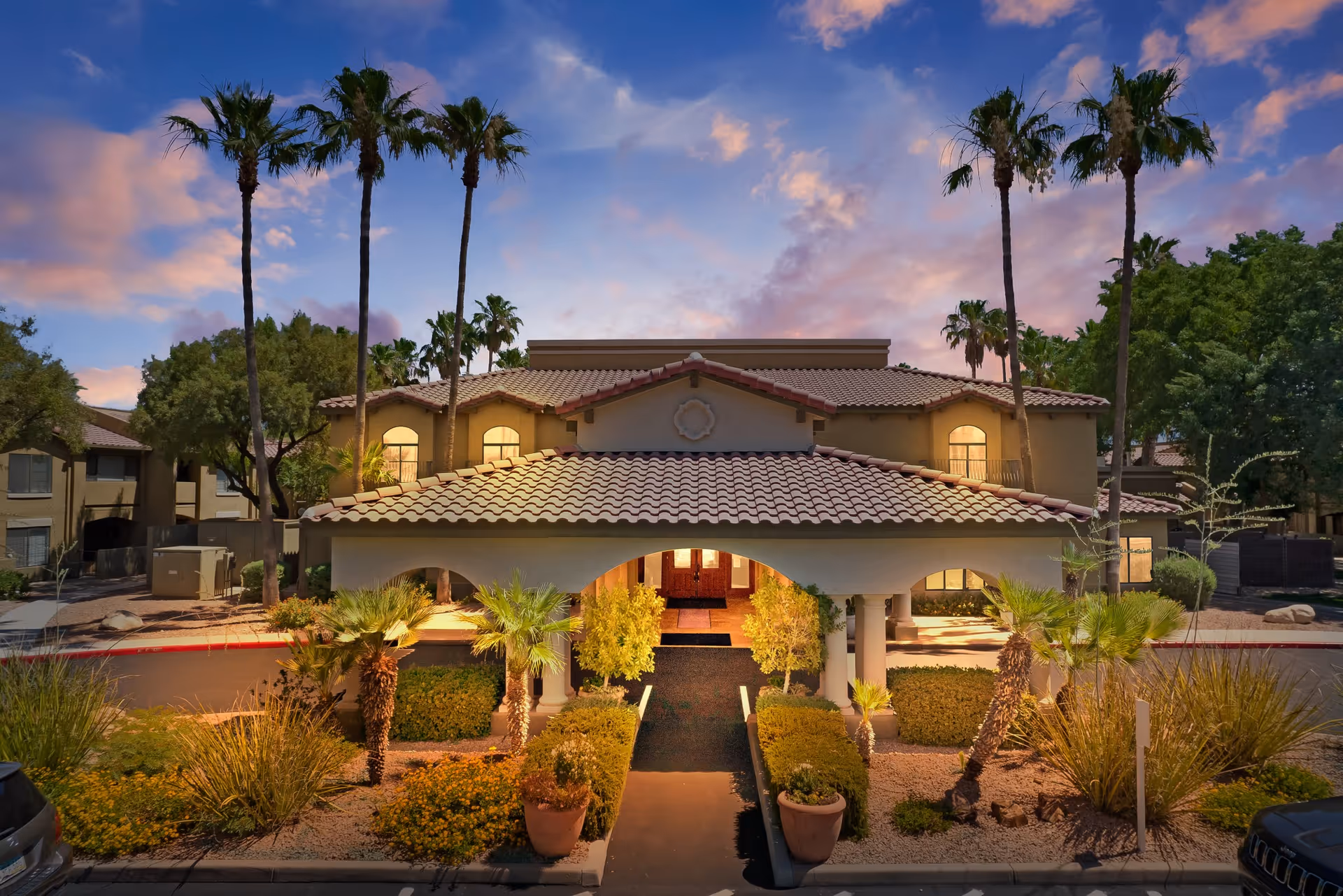 Exterior view of a two-story building with a tiled roof and an arched entrance, surrounded by palm trees and desert landscaping under a colorful sunset sky.