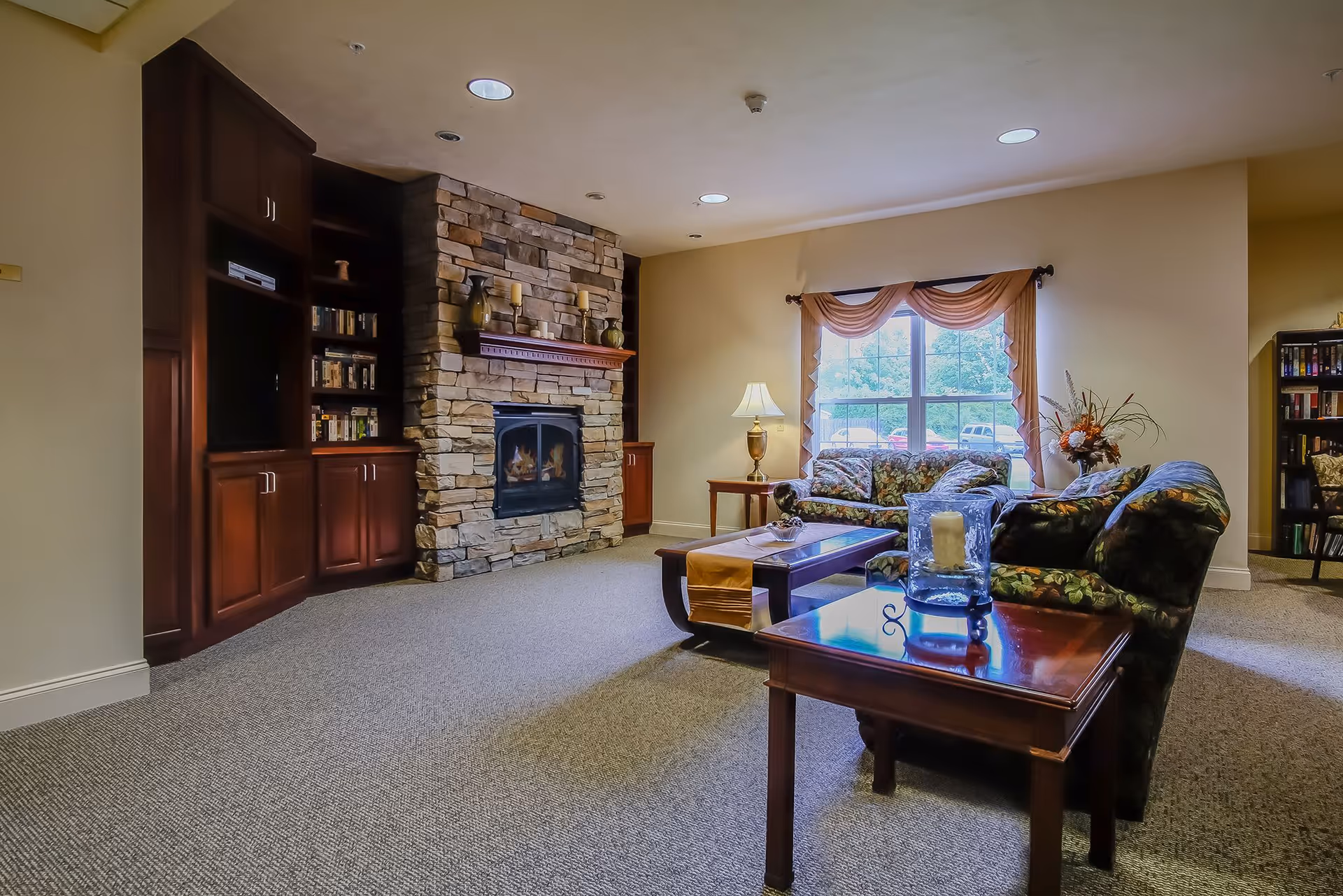 A cozy living room with a stone fireplace surrounded by built-in wooden shelves and cabinets. There are two floral-patterned sofas, a wooden coffee table with a decorative runner, and a side table with a lamp and a candle holder. A large window with draped curtains lets in natural light, and there is a vase with flowers near the window.