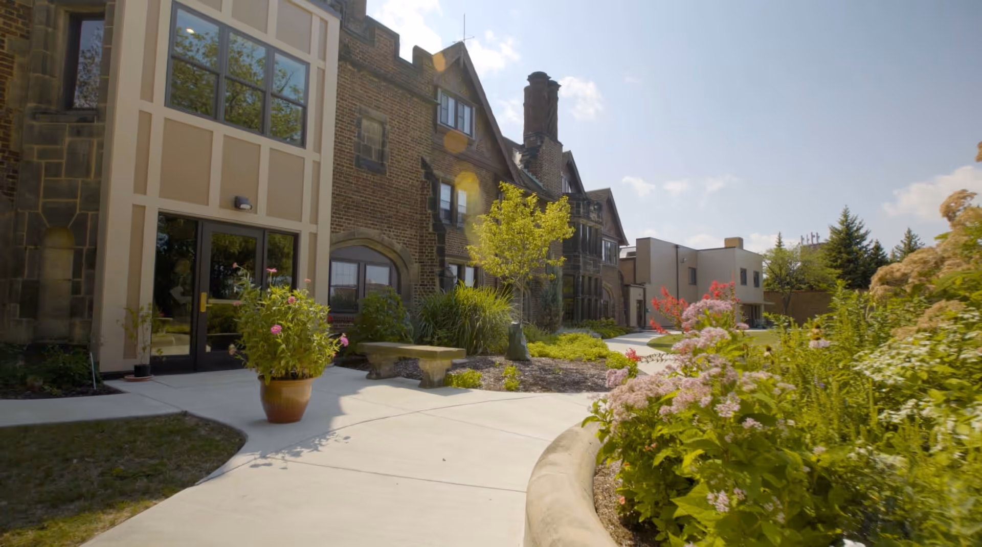 Outdoor view of a senior living community building with a stone and brick facade, large windows, and a pathway surrounded by green plants and colorful flowers under a clear sky.