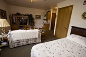 A cozy senior living facility room featuring a bed with floral bedding in the foreground, a floral-patterned sofa with a white throw, a wooden entertainment center with a TV and decorative items, a small kitchen area with cabinets and appliances, and a wooden door. The room has neutral-colored walls and carpeted flooring.