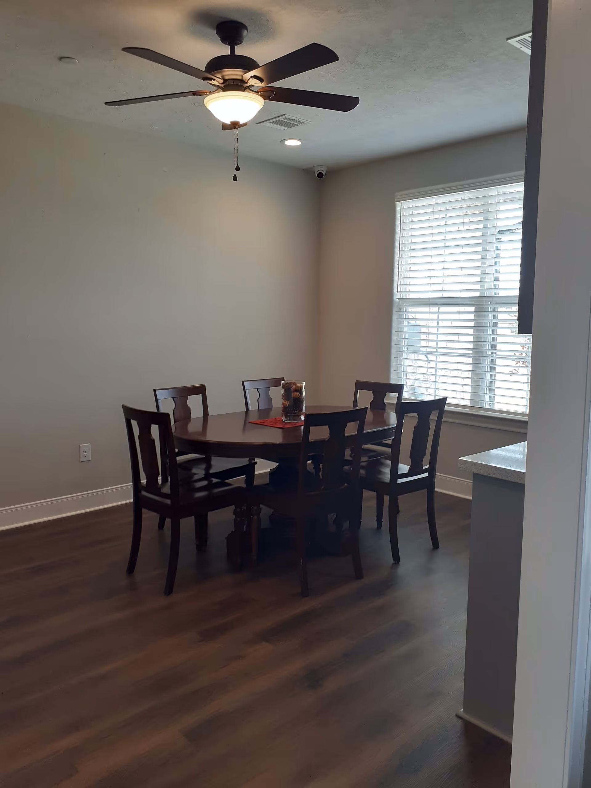 A dining room with a dark wood table and six chairs under a ceiling fan beside a window with blinds.