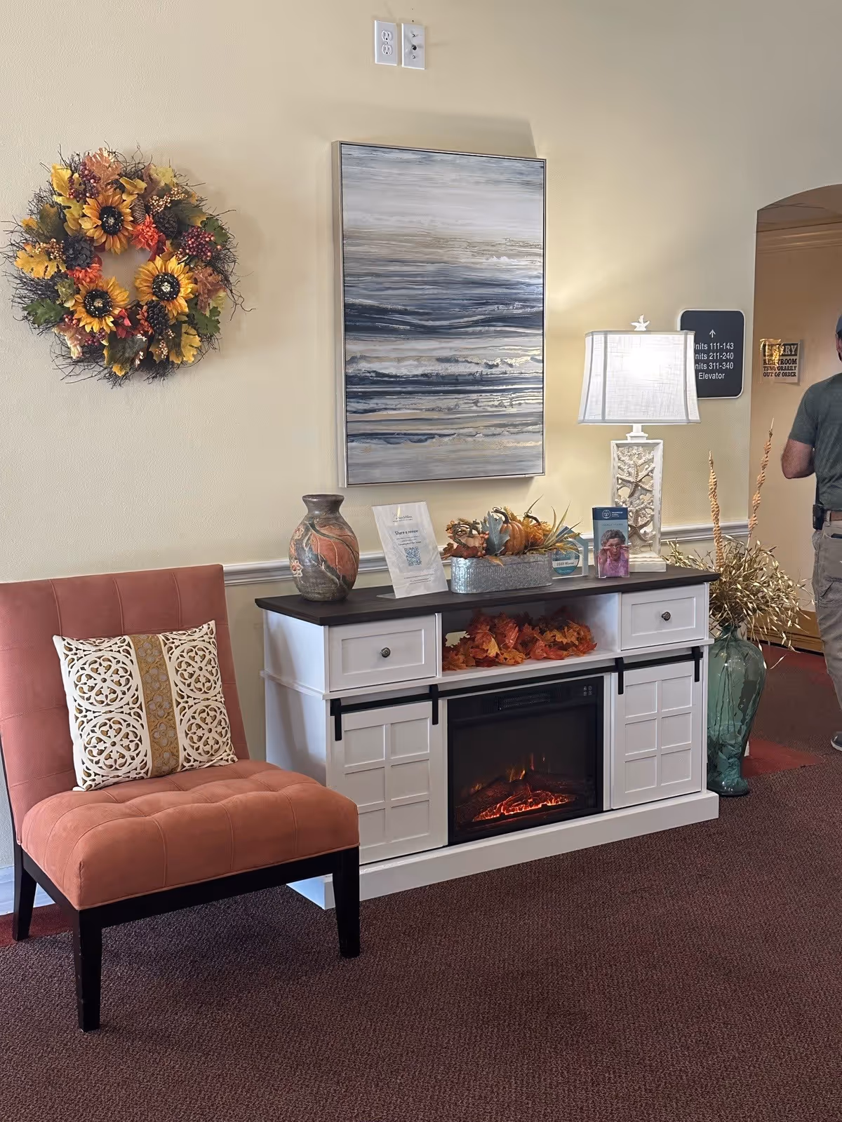 A cozy corner in a senior living facility featuring a coral upholstered chair with two decorative pillows, a white cabinet with a built-in electric fireplace, and autumn-themed decorations including a wreath with sunflowers on the wall and a centerpiece on the cabinet. A modern abstract painting hangs above the cabinet, and a lamp with a white shade is placed on top. A person is partially visible walking through an archway in the background.