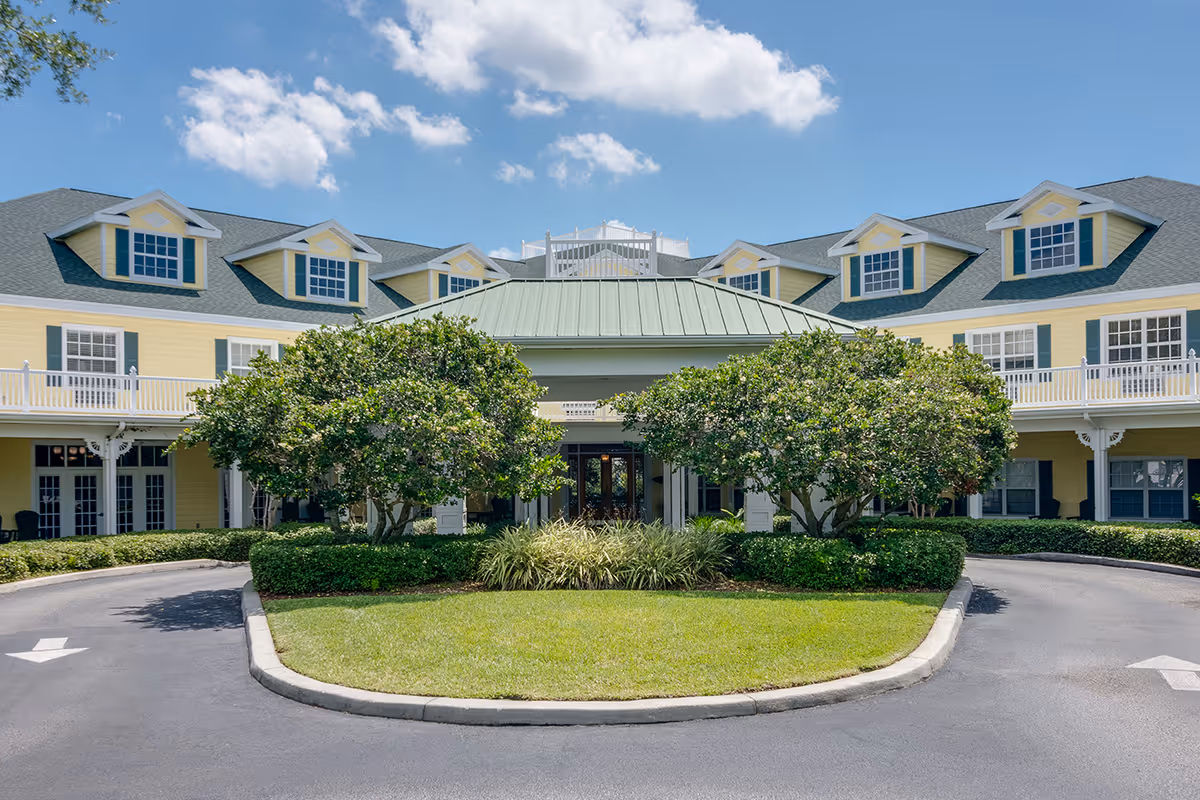 Front exterior view of a senior living facility named Arbor Oaks at Tyrone, featuring a yellow two-story building with white trim, green roof, multiple dormer windows, and a circular driveway with landscaped greenery and trees in the center.
