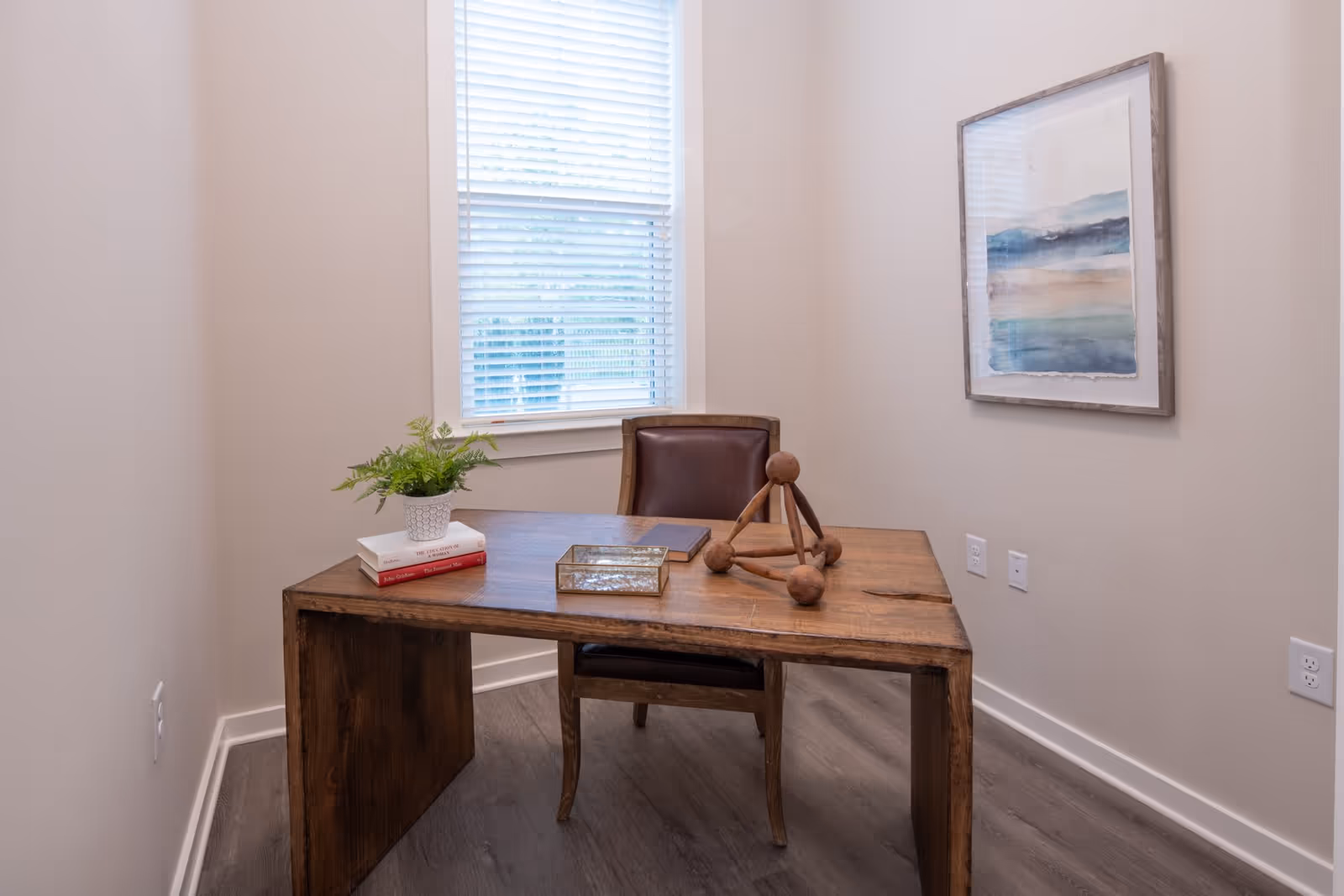 A small office or study room with a wooden desk and a brown leather chair. On the desk are a small potted plant, two stacked books, a closed book, a decorative wooden geometric sculpture, and a glass box. A window with white blinds is behind the desk, and a framed abstract painting with blue and beige tones hangs on the wall to the right.