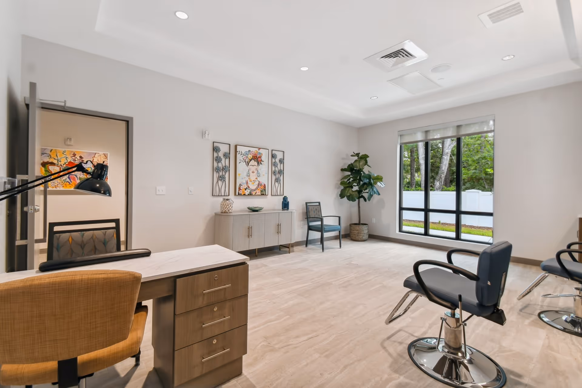 Bright modern interior room with salon chairs, a desk, console table with artwork, and a large window overlooking trees.