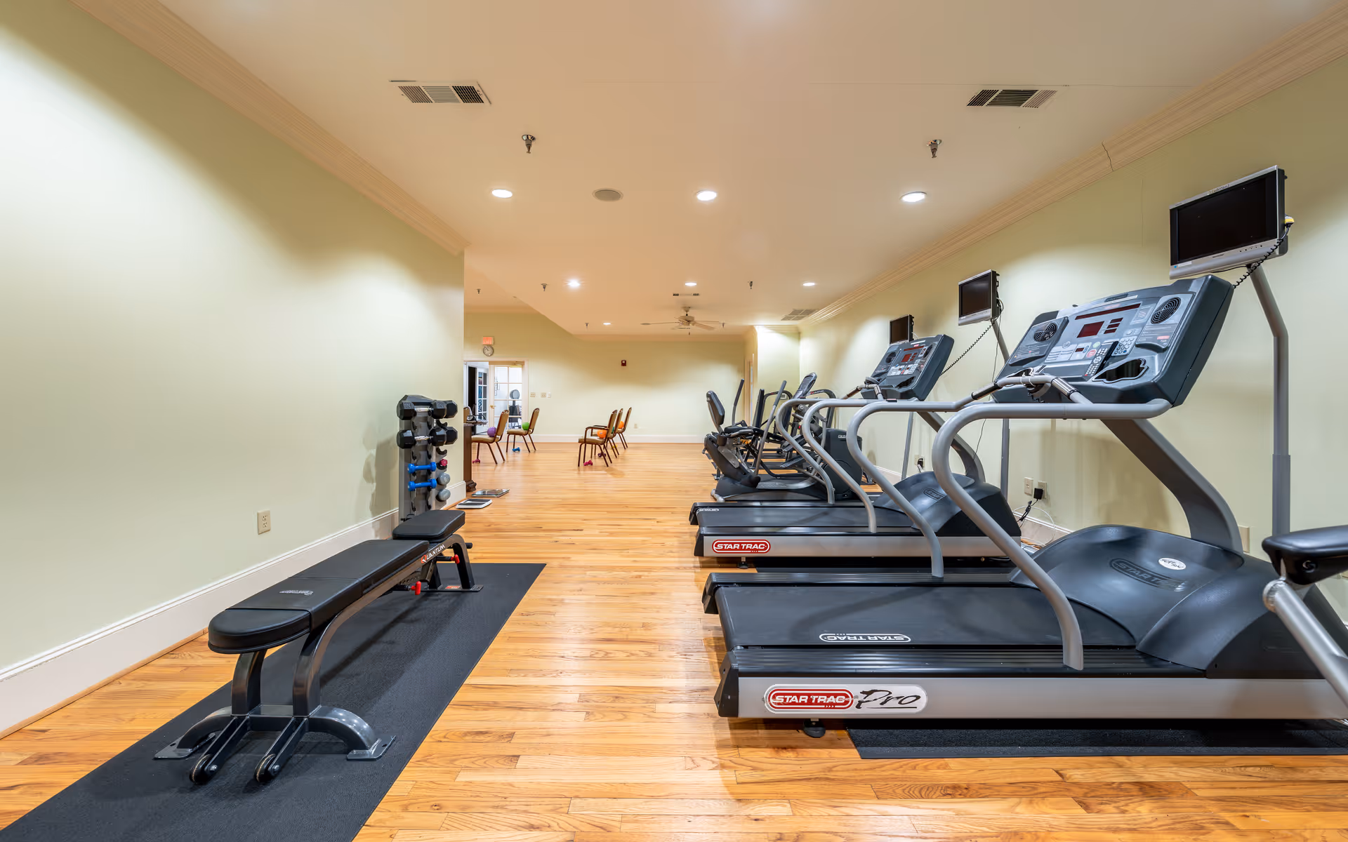 Interior view of a fitness room with wooden flooring, featuring three treadmills with attached screens on the right side and a weight bench with dumbbells on the left side. In the background, there are a few chairs and a table near a door.