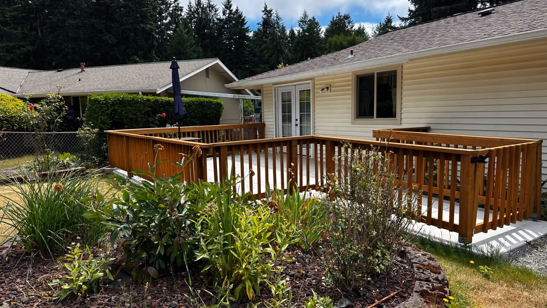 Outdoor patio area with wooden railing attached to a beige single-story building. There is a garden with various green plants and flowers in the foreground, and another building with a chain-link fence and greenery in the background under a partly cloudy sky.