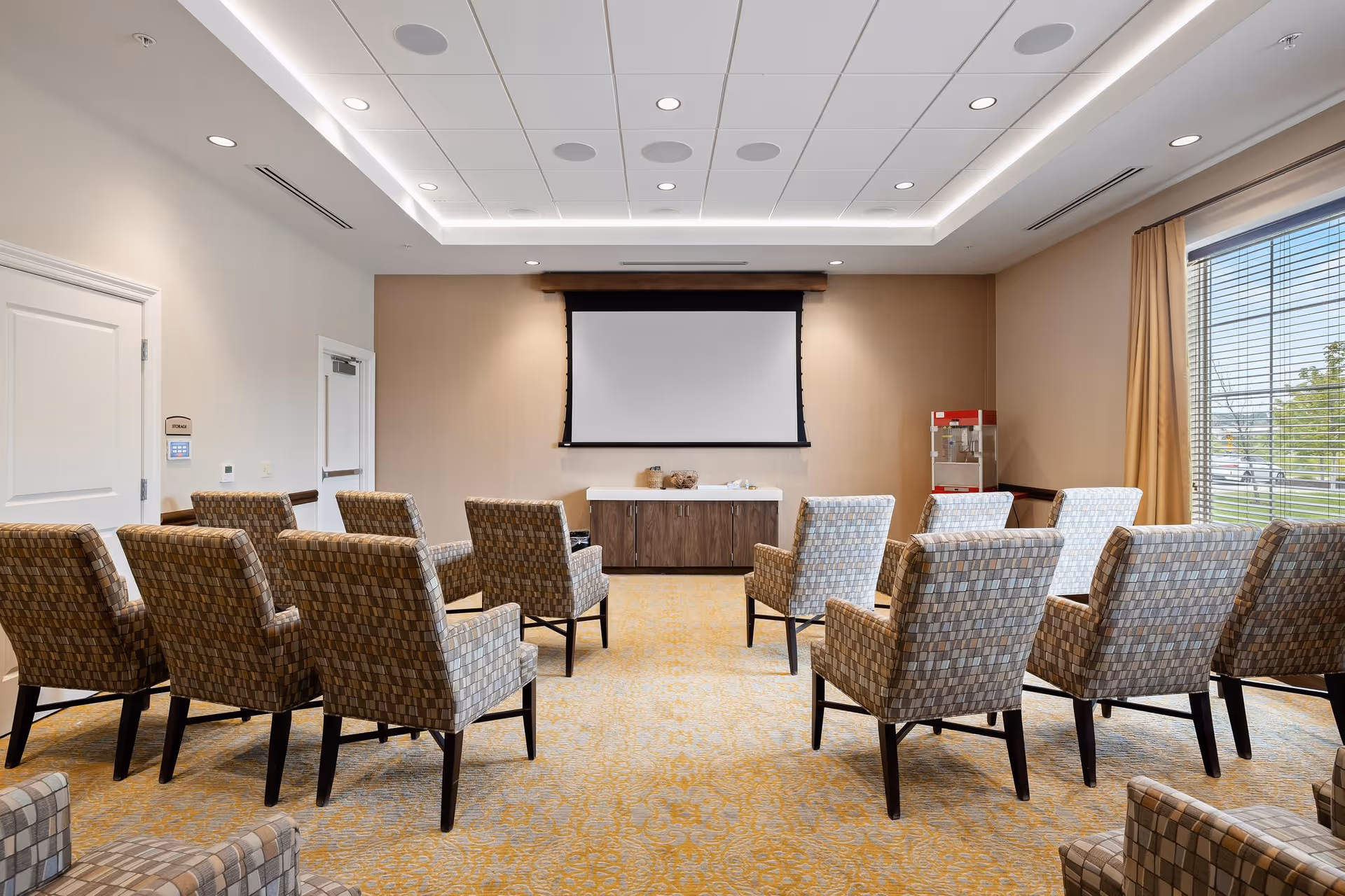 A small theater or presentation room with rows of patterned armchairs facing a blank projection screen mounted on a beige wall. There is a cabinet below the screen and a popcorn machine in the corner near a large window with beige curtains. The room has a carpeted floor and a white ceiling with recessed lighting.