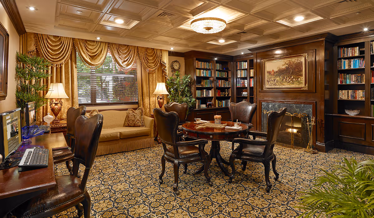 A cozy, elegant living room with a patterned carpet, a round wooden table surrounded by four leather chairs, a beige sofa with decorative pillows, two table lamps with ornate bases, large windows with gold drapes, built-in wooden bookshelves filled with books, a fireplace with a painting above it, and a computer workstation with a keyboard and monitor on the left side.