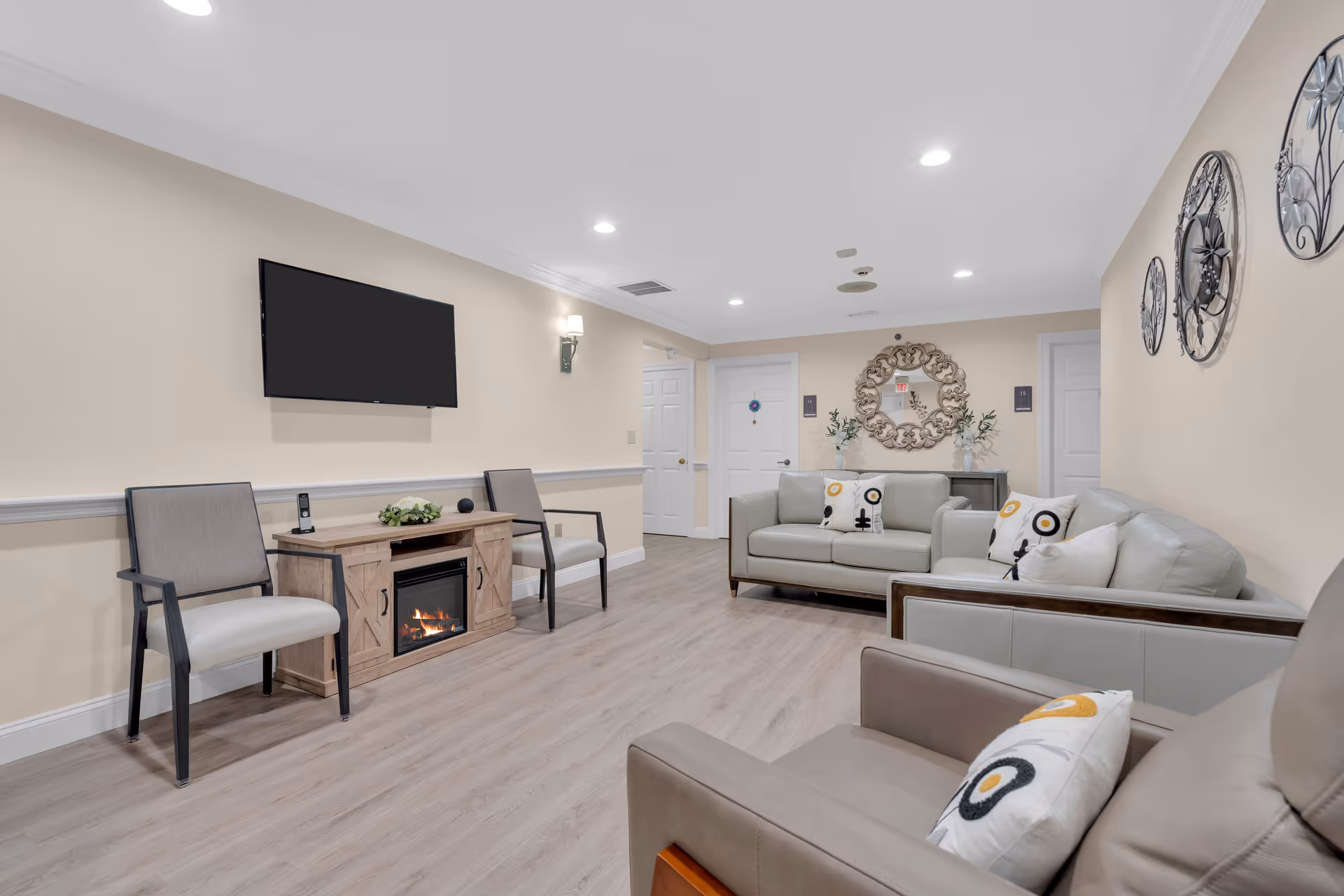 A cozy living room with beige walls and light wood flooring. The room features a wall-mounted flat-screen TV above a wooden cabinet with a built-in electric fireplace. There are two gray armchairs with black frames on one side and two light gray sofas with decorative pillows on the other side. A large decorative mirror and plants are placed on a console table against the far wall. The room is well-lit with recessed ceiling lights and wall sconces.