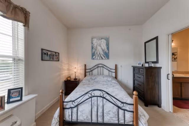 A small bedroom with a single bed featuring a metal and wood frame, a nightstand with a lamp, a dresser with a mirror, and a window with blinds and a valance. There is artwork on the walls and a doorway leading to a bathroom.