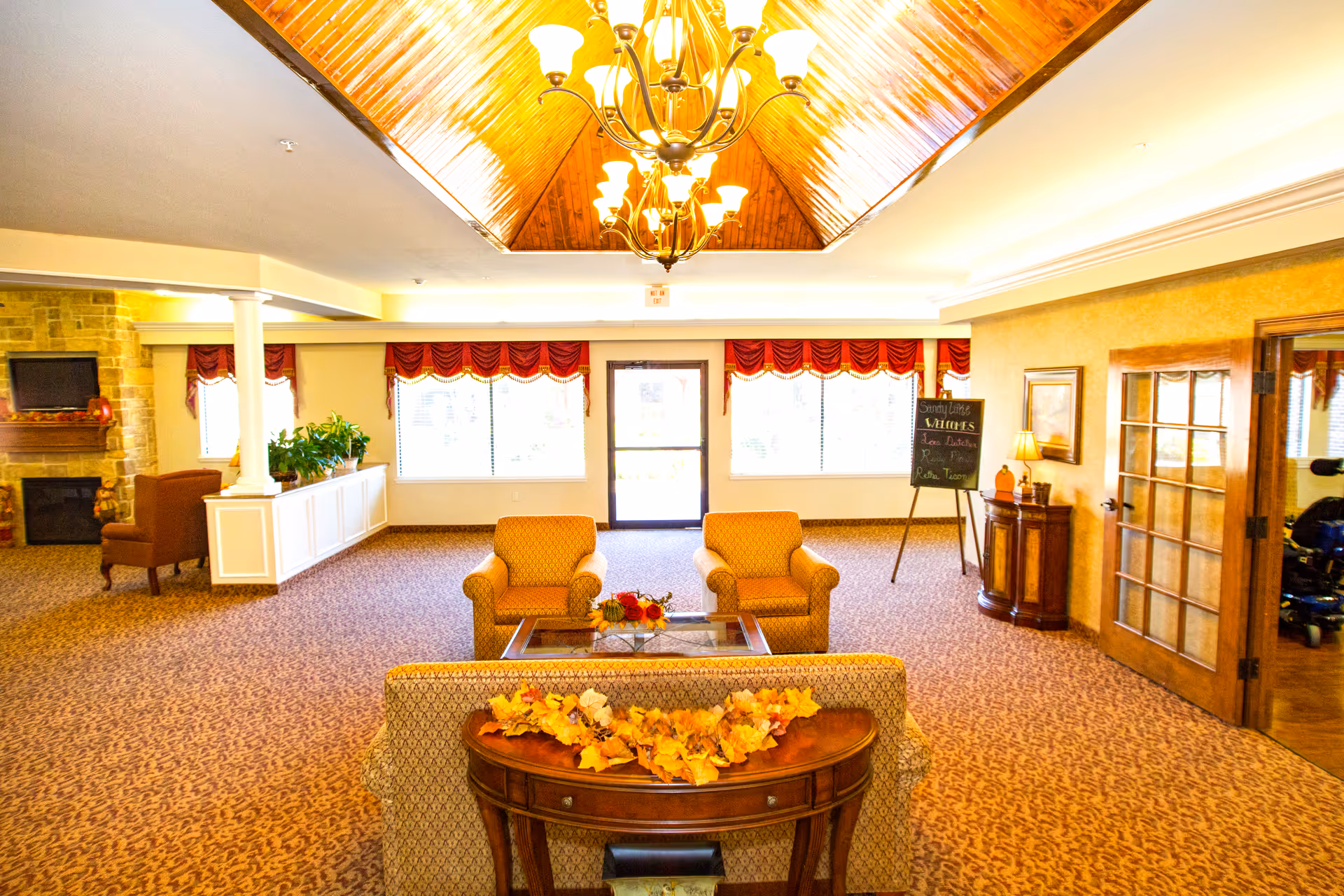 A spacious and warmly lit living room area with patterned carpet, two armchairs, a sofa with a wooden console table decorated with autumn leaves, a glass coffee table with a floral centerpiece, and a stone fireplace with a TV mounted above it. The ceiling features a wooden vaulted design with chandeliers, and large windows with red valances allow natural light to fill the room. A chalkboard sign and wooden furniture are visible near a glass-paneled door.