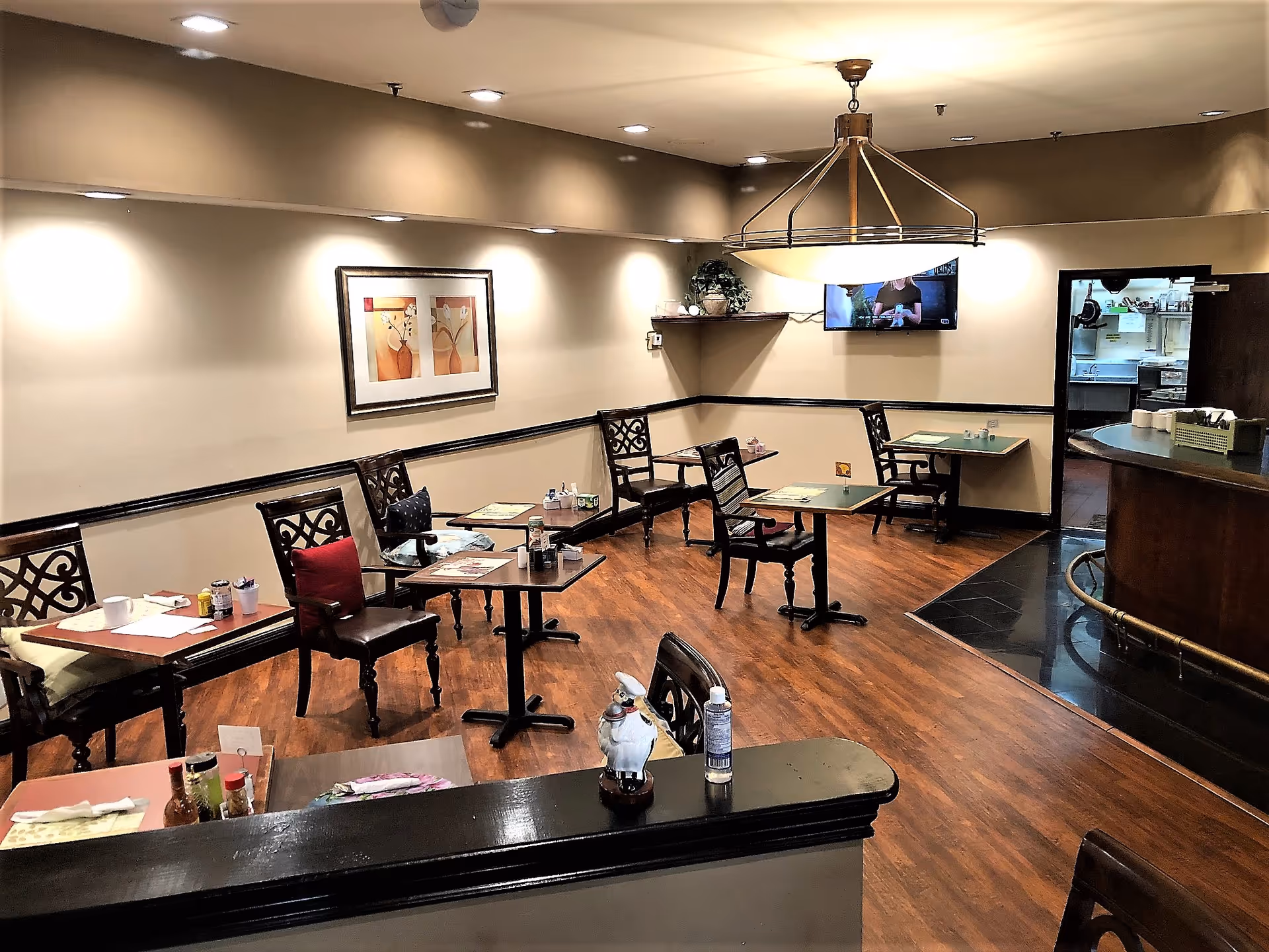 A cozy dining area in a senior living facility with several small tables and chairs arranged along the walls. The room has warm lighting, wooden flooring, a framed artwork on the wall, a flat-screen TV mounted on the wall, and a counter area to the right. The space appears clean and inviting with some condiments and napkins on the tables.
