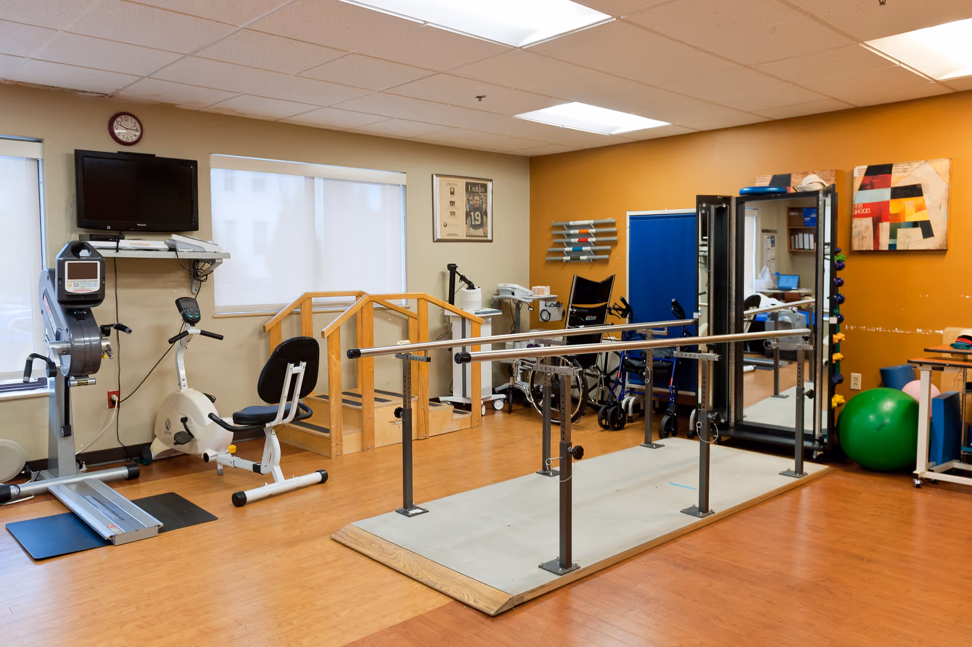 A physical therapy or rehabilitation room with exercise equipment including a stationary bike, parallel bars for walking practice, a small wooden staircase for step exercises, a large mirror, and various therapy tools such as exercise balls and weights. The room has wood flooring, beige and orange walls, and a wall-mounted TV.