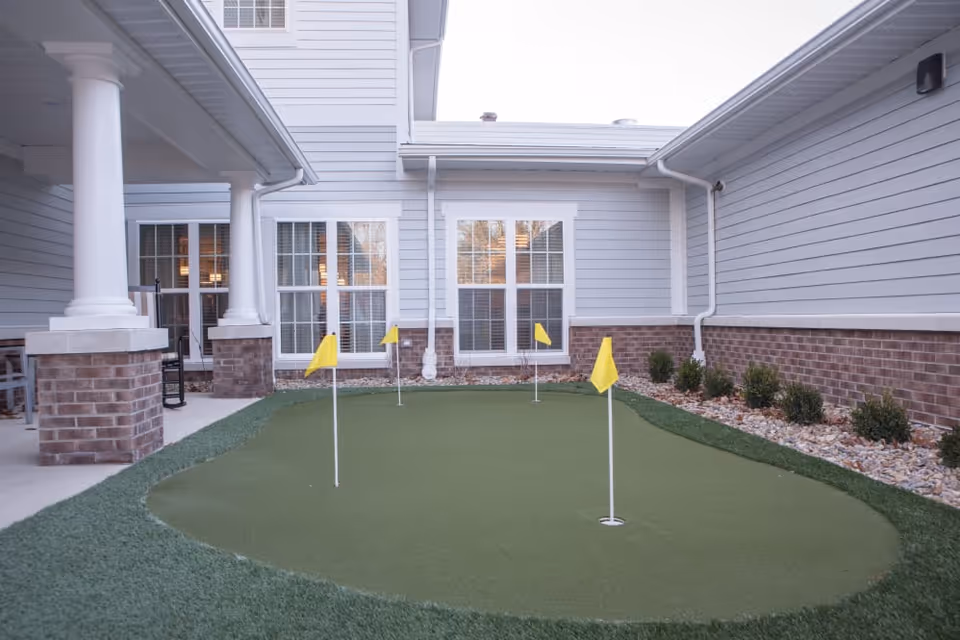 Small outdoor putting green with four yellow flags surrounded by a building with white siding and brick accents. There are windows and a covered patio area with white columns on the left side.
