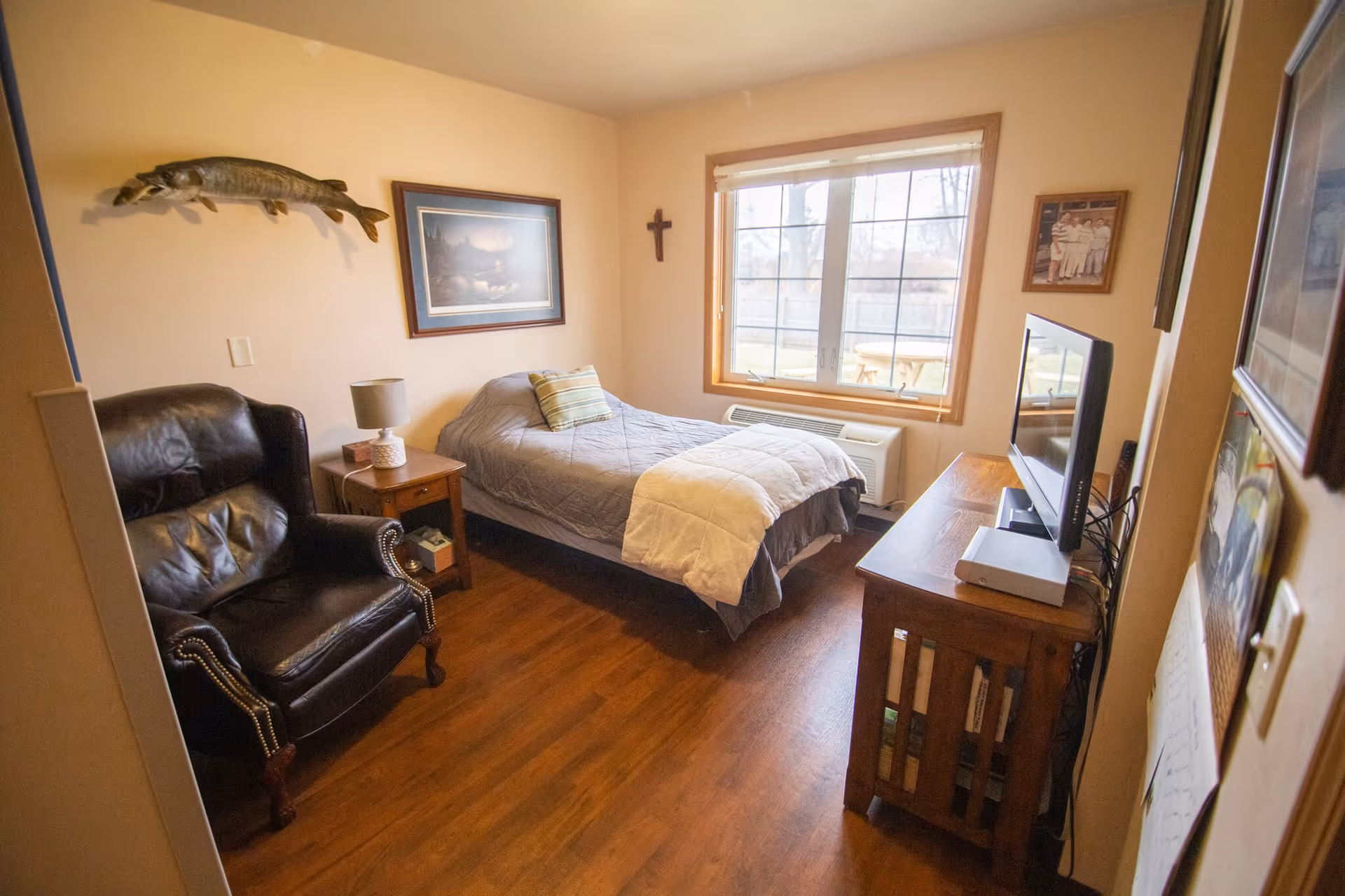 A cozy bedroom with a single bed covered in a gray quilt and a white comforter. There is a black leather armchair with nailhead trim, a wooden nightstand with a lamp, and a wooden dresser with a flat-screen TV on top. The room has wooden flooring, a large window with a view of an outdoor patio, and wall decorations including a mounted fish, framed pictures, and a cross.